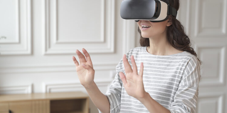 Young impressed brunette businesswoman interacting with virtual reality wearing VR headset glasses while relaxing between work online from home. Exited happy lady playing 3D video games in cyberspace